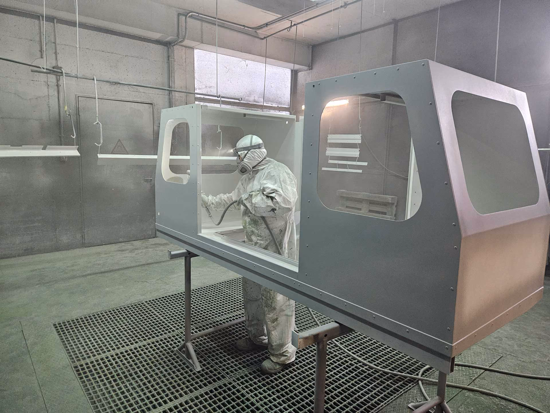 Person in Schutzkleidung lackiert ein grosses Metallgehäuse in einer Werkstatt.