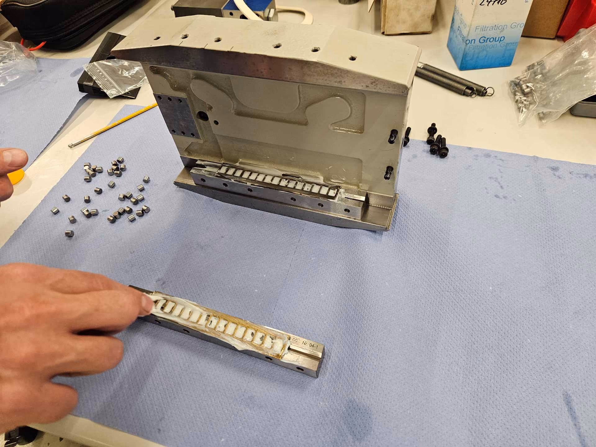 Hand working on a metal mold with small cylindrical parts on a blue mat on the workbench.