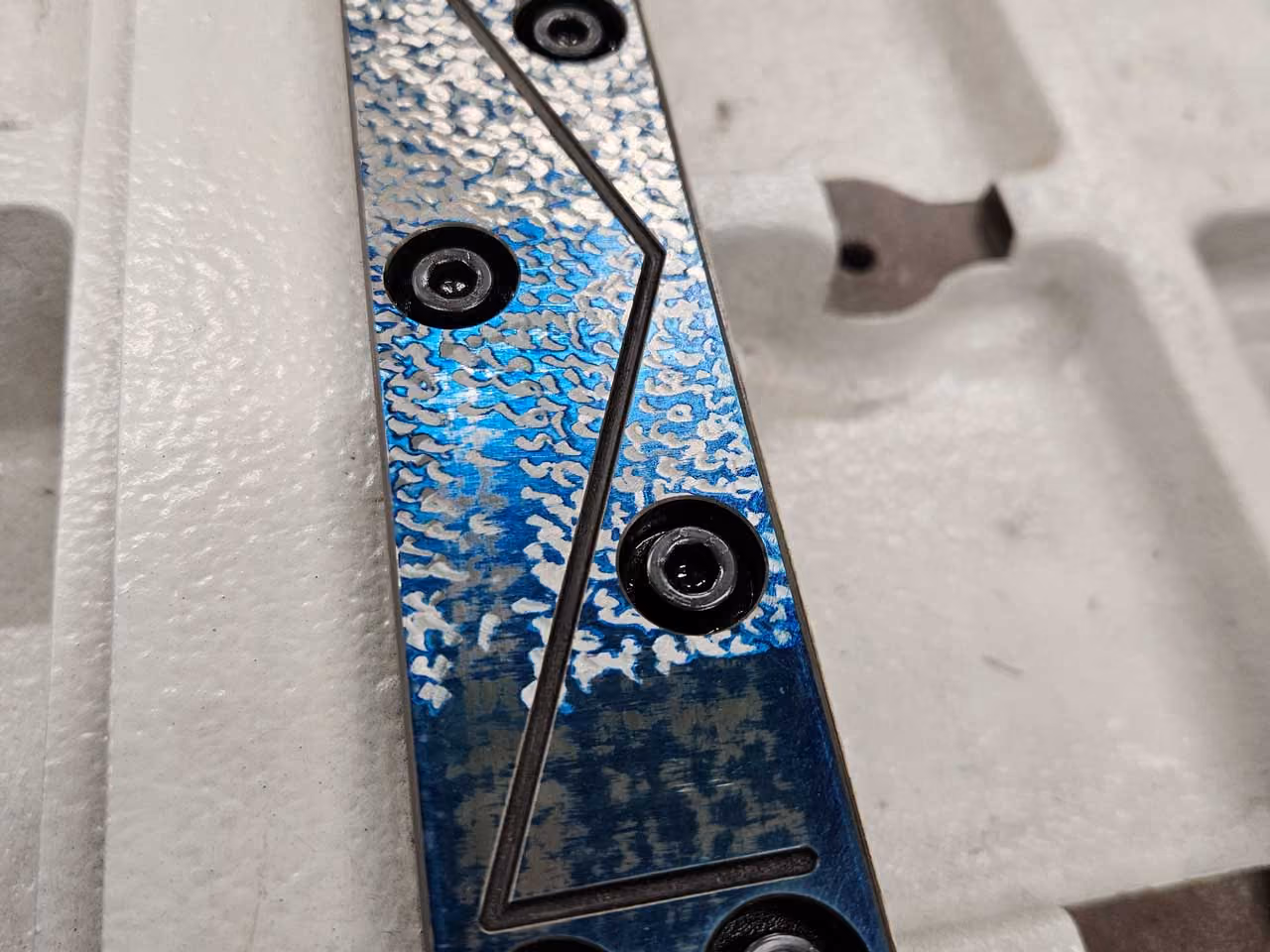 Blaues Metallteil mit silbernen Mustern und schwarzen Schrauben auf weißem Schaumstoff.