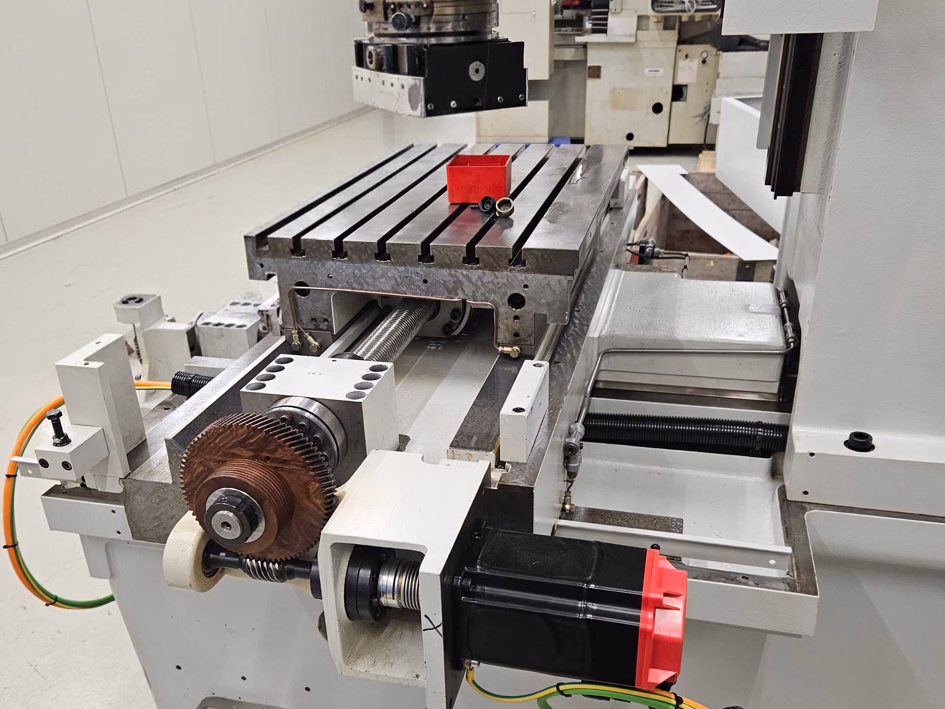 Präzisionsmaschine mit Metall-Tisch und Schraubspindel, davor rotes Werkzeugteil.