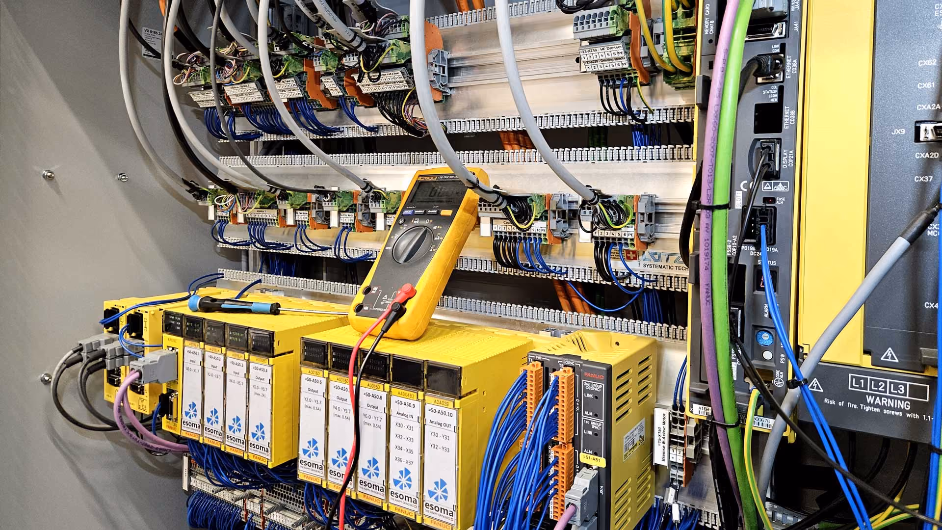 Elektronisches Steuerungssystem mit gelben Esoma-Fanuc-Modulen und gelbem Multimeter zur Messung.