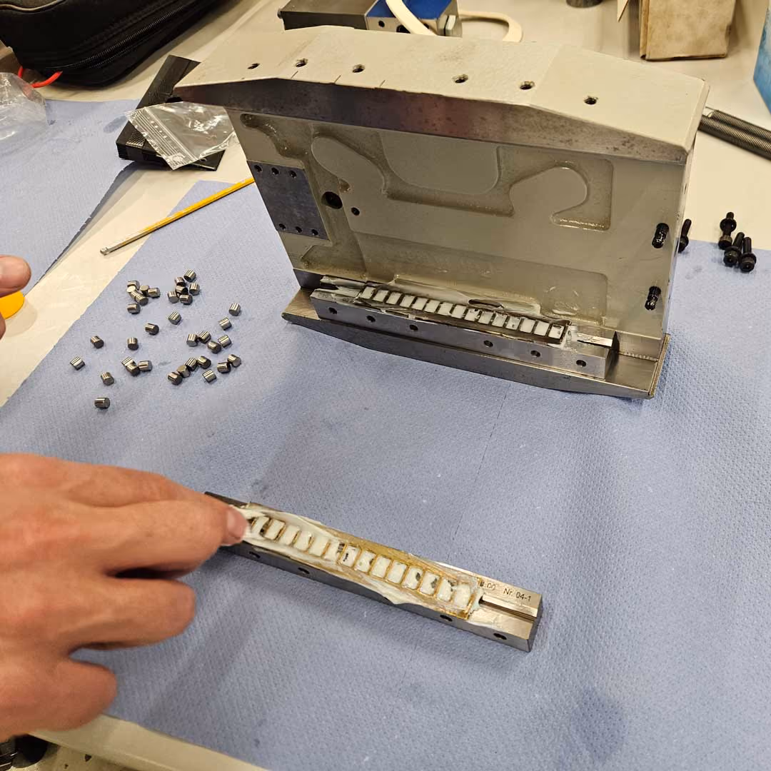 Hand holding a metal mold piece near a larger mold on a blue cloth with small cylindrical metal parts nearby.