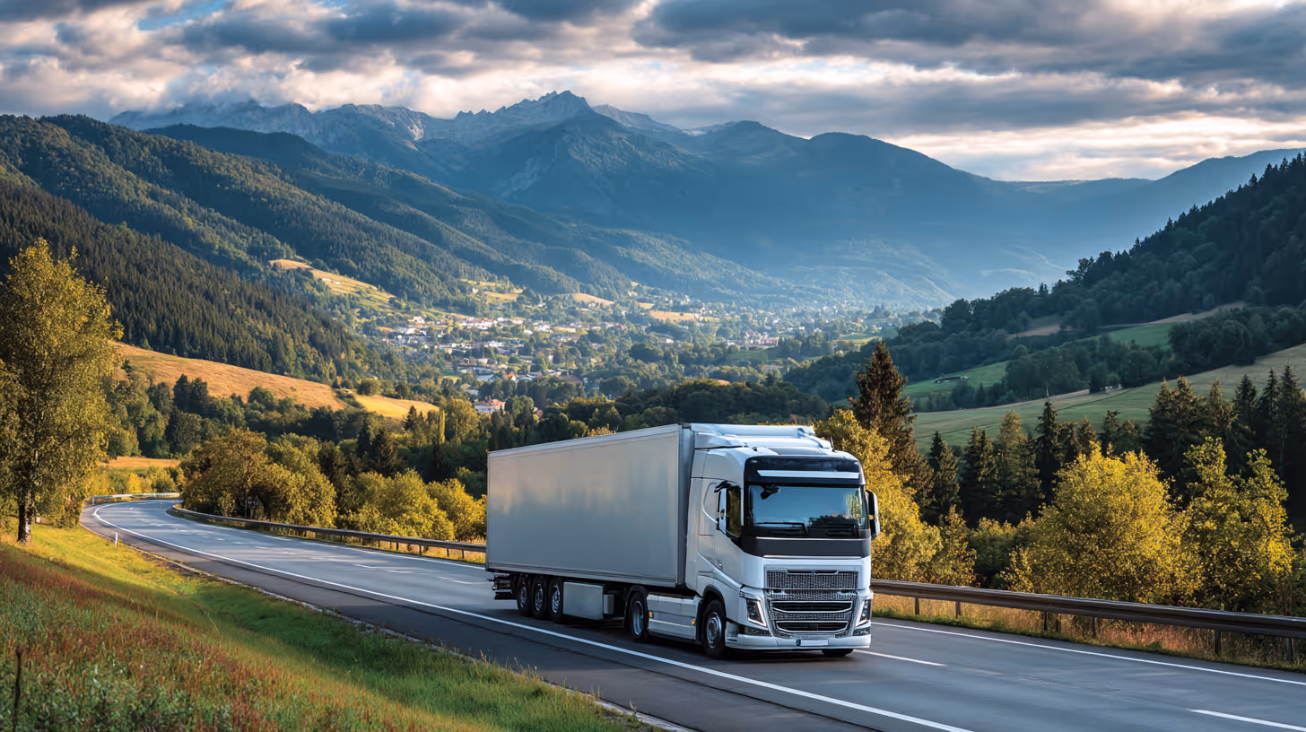 White semi-truck driving on a highway surrounded by green hills and mountains under a cloudy sky.