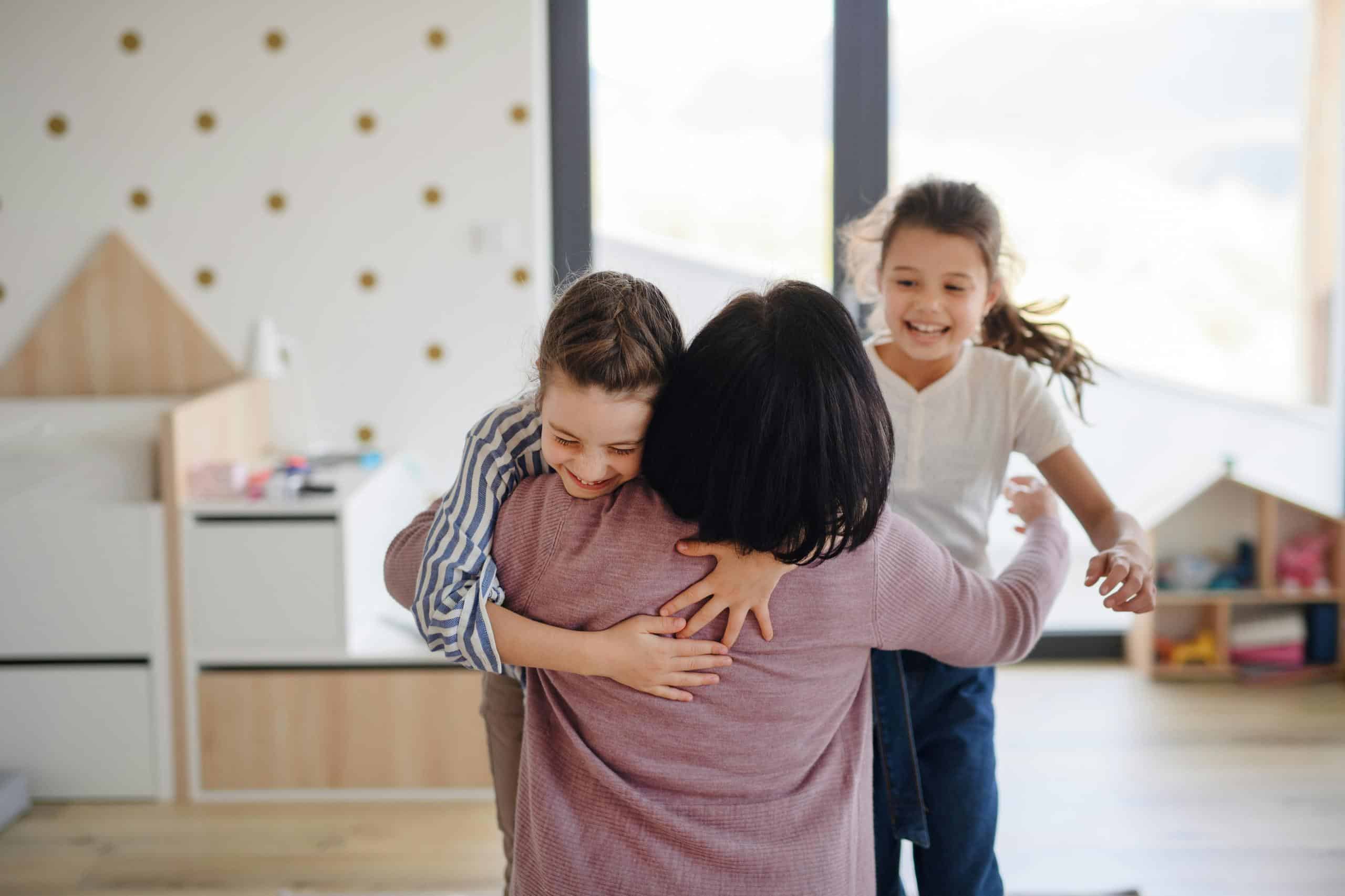 Caregiver kneeling to embrace one child while another joins in, showing inclusive caregiving through responsive attention, repair, and connection during daily routines.