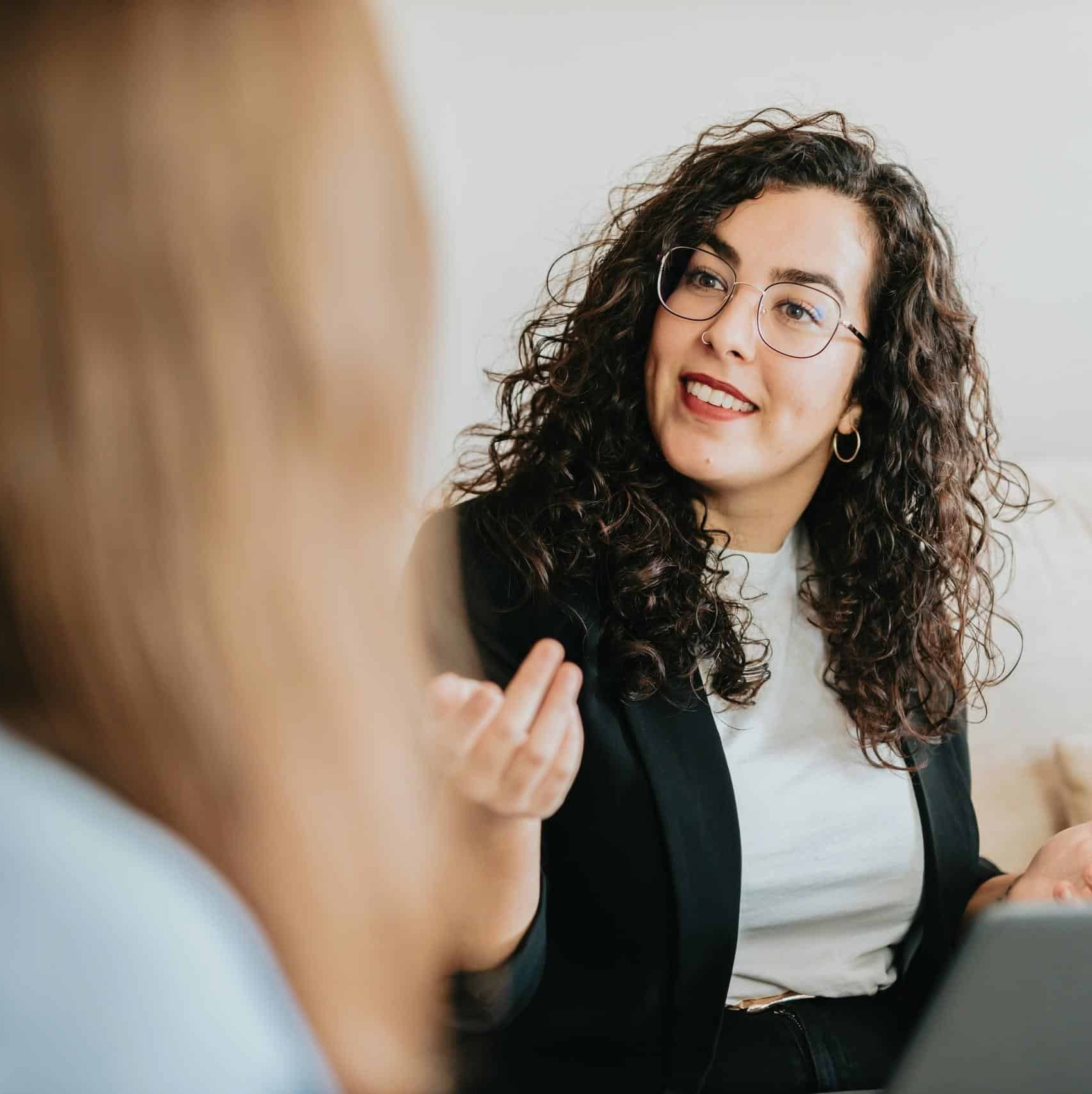 A woman with curly hair and glasses speaks confidently during a professional conversation, gesturing with her hand