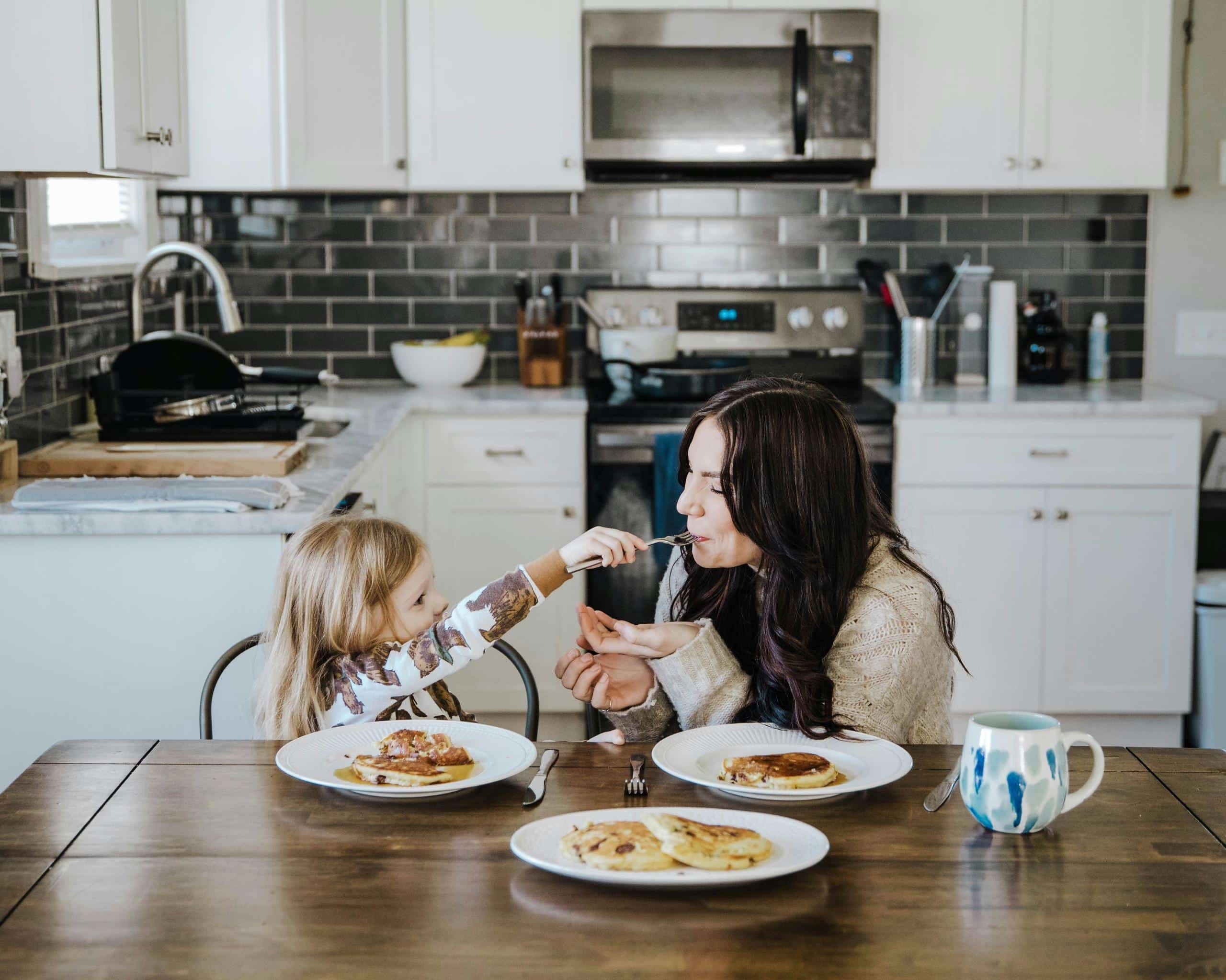 A caregiver with long dark hair sits at a kitchen table sharing a meal with a young blonde girl, both leaning in toward each other in a warm and attentive moment