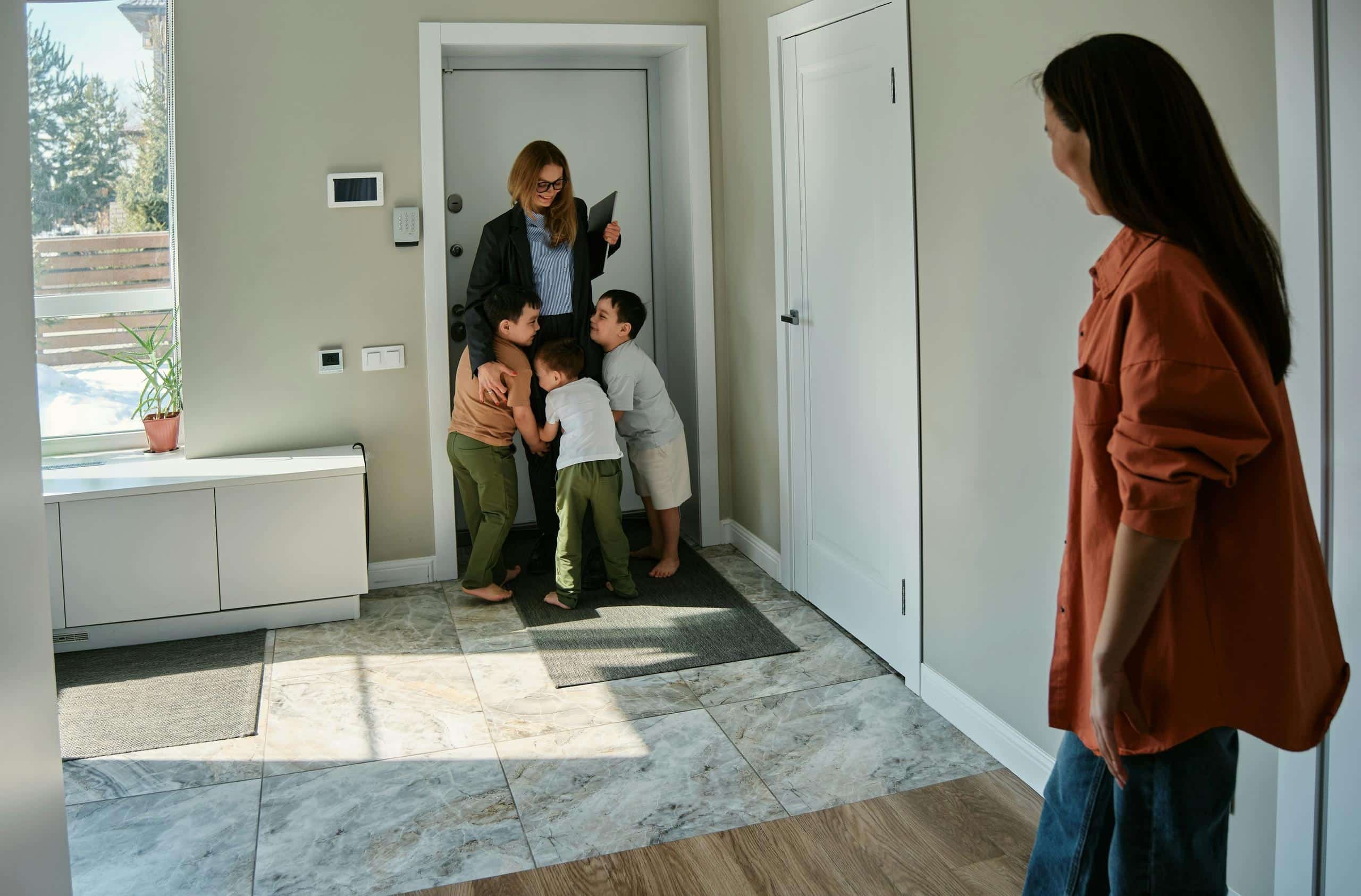 A woman in a rust-colored shirt watches from across an entryway as two young children greet a caregiver arriving at the front door