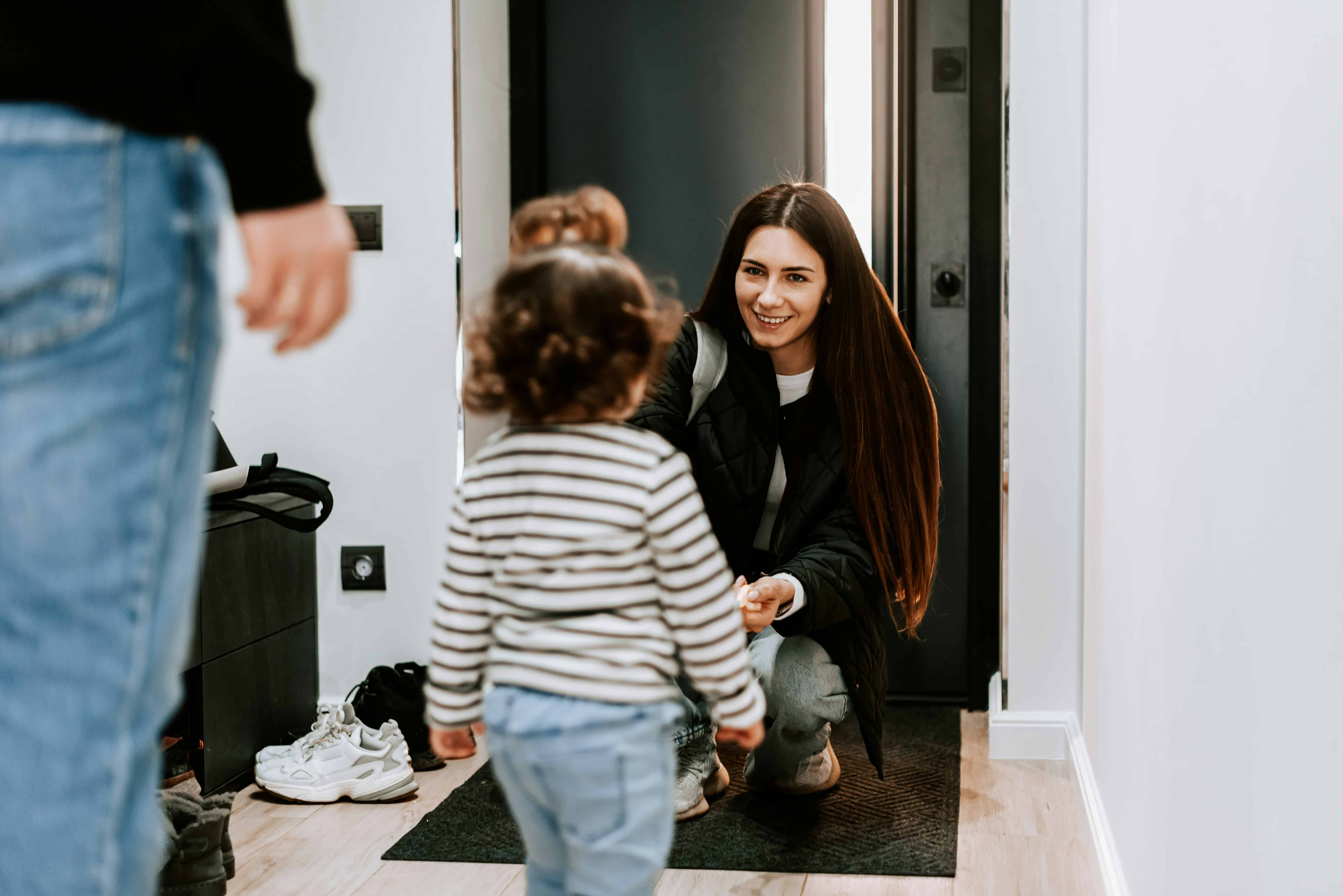 A nanny crouching down to greet a toddler at the front door as a parent arrives home