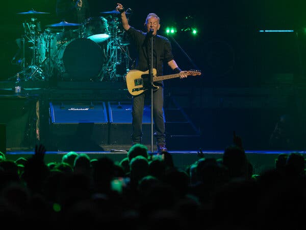 Bruce Springsteen, dressed in black with his Telecaster, raises an arm onstage in front of a crowd.