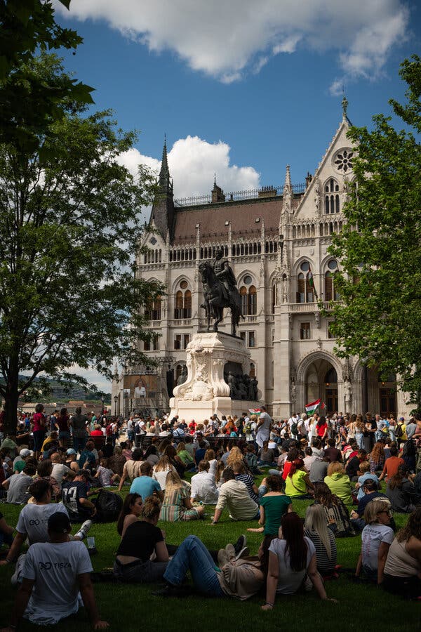 Assorted scenes of happy Hungarians celebrating on a beautifully sunny day — sitting on a lawn and watching a ceremony, clustered around a bronze statue of a seated man, and dancing near the river.