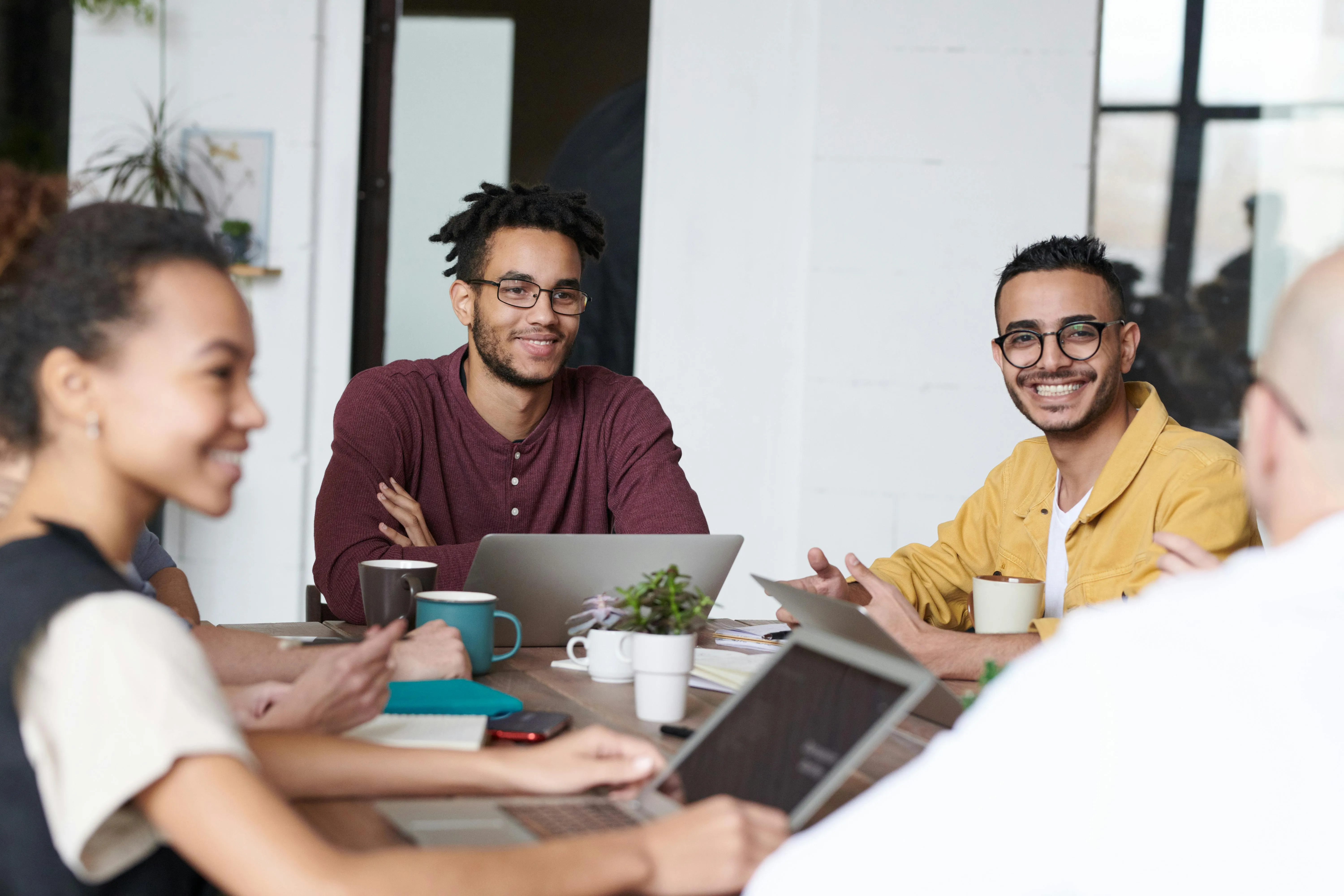 Group of diverse young adults smiling and discussing ideas around a table with laptops and coffee cups in a modern office.
