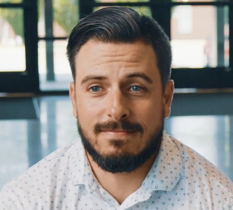 Close-up of a bearded man with blue eyes wearing a white patterned shirt, seated indoors with large windows in the background.