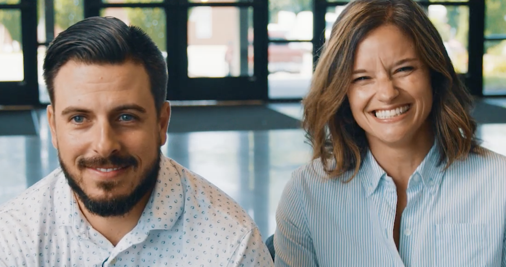 Smiling man with a beard and woman with shoulder-length hair, both wearing light-colored shirts, seated indoors near glass doors.
