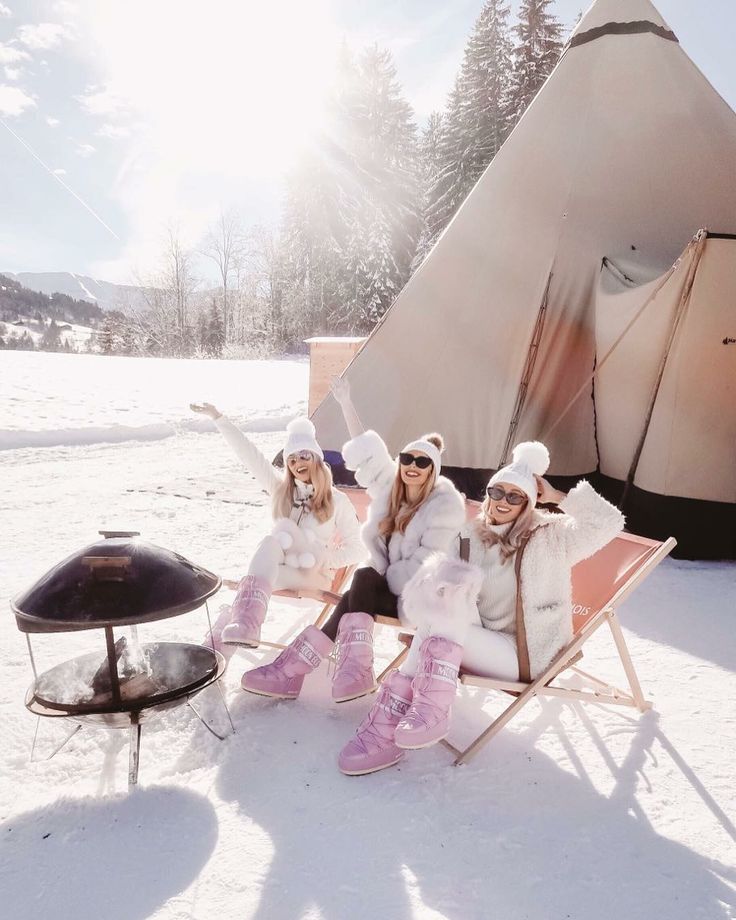 Three women wearing white winter outfits and pink boots sitting in deck chairs in snow next to a teepee and fire pit, smiling and waving.