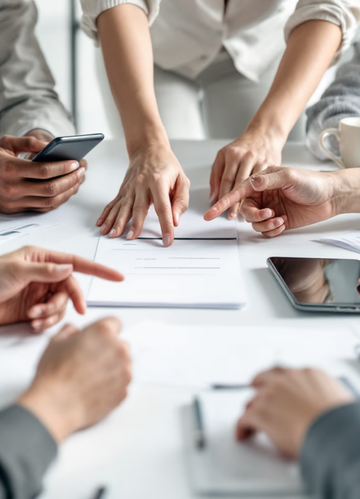 People at a table pointing and discussing documents with smartphones and notebooks nearby.