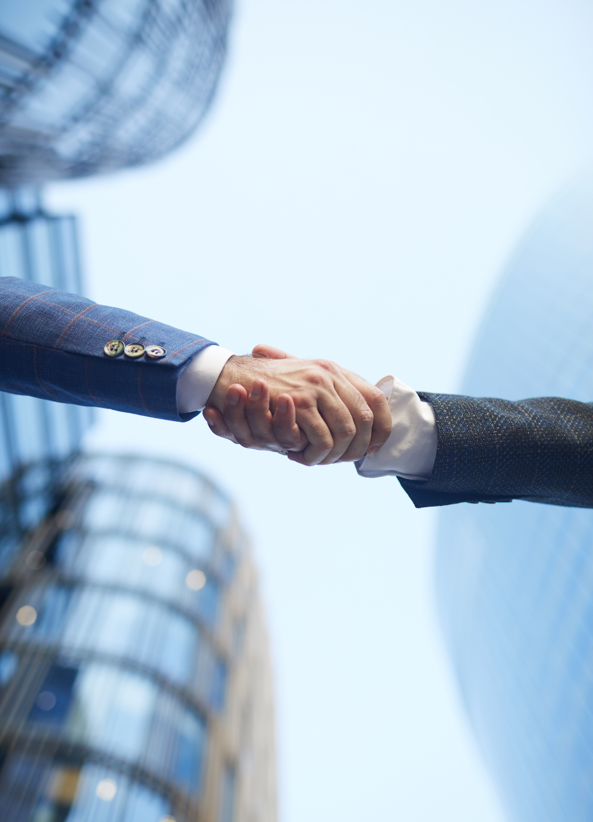 Two people in suits shaking hands outdoors with modern glass buildings in the background.