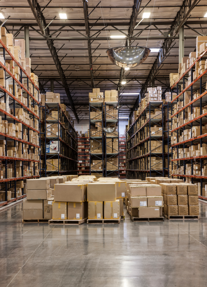 Warehouse aisle with tall shelves and pallets stacked with cardboard boxes on a shiny concrete floor.