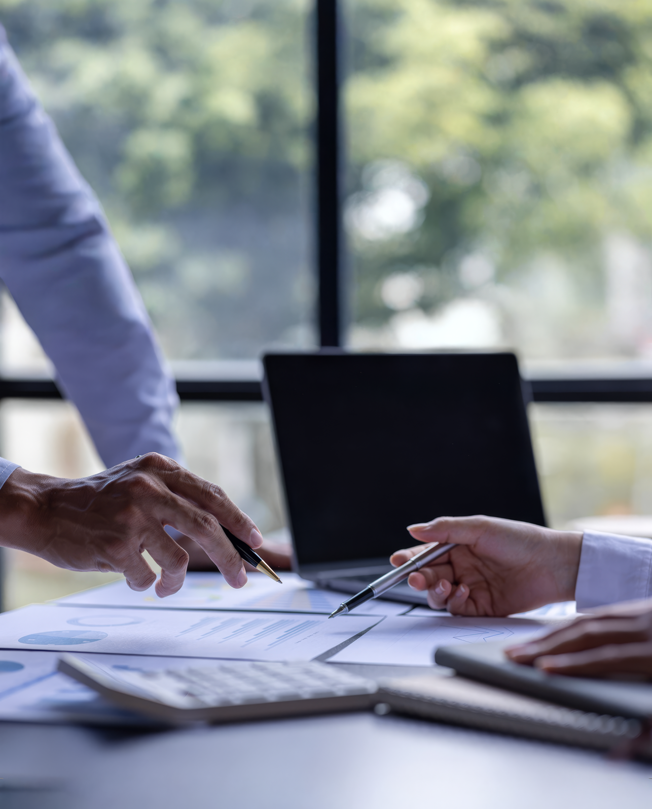 Two people pointing at documents on a desk with pens, a calculator, and a laptop in the background.
