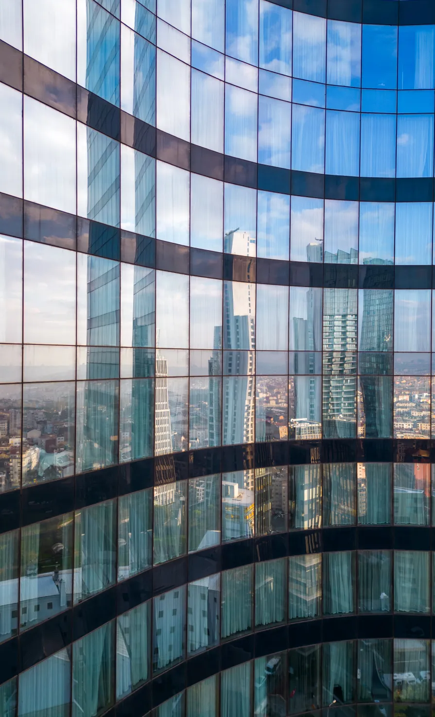 Curved glass building facade reflecting tall skyscrapers and a blue sky with clouds.