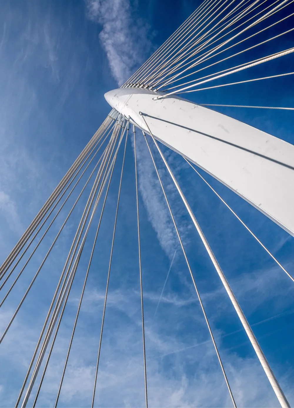 Upward view of a white cable-stayed bridge tower against a blue sky with wispy clouds.