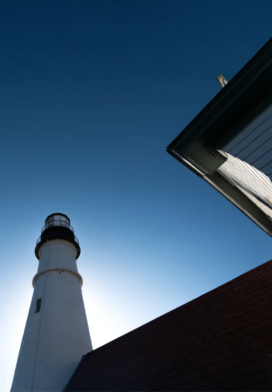 White lighthouse with black top beside building roofs under clear blue sky, sun behind lighthouse.