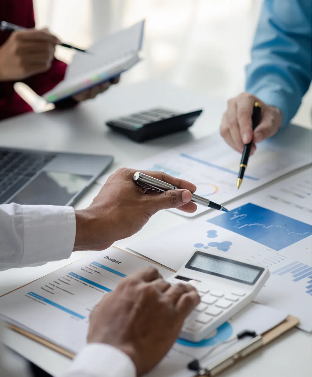 Hands using a calculator and pointing at financial documents with charts and a budget sheet on a desk.