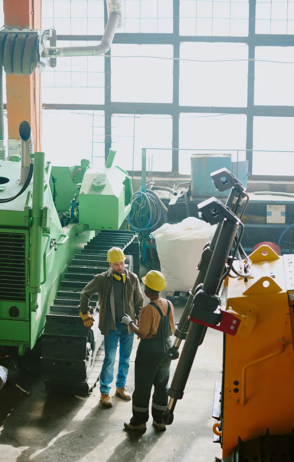Two factory workers in hard hats talking next to large industrial machines in a workshop.