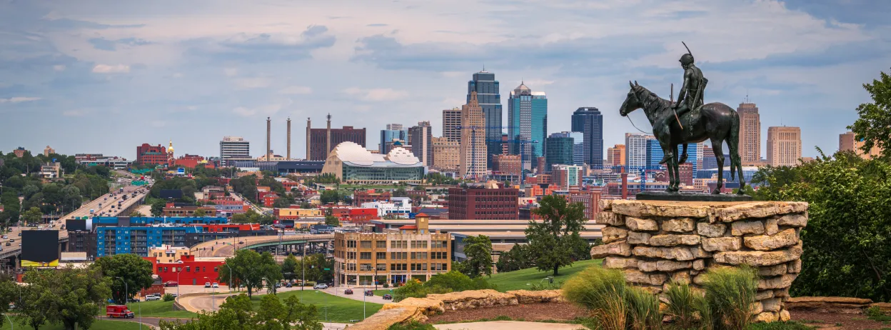 Statue of a Native American on horseback overlooking Kansas City skyline with skyscrapers and cloudy sky.