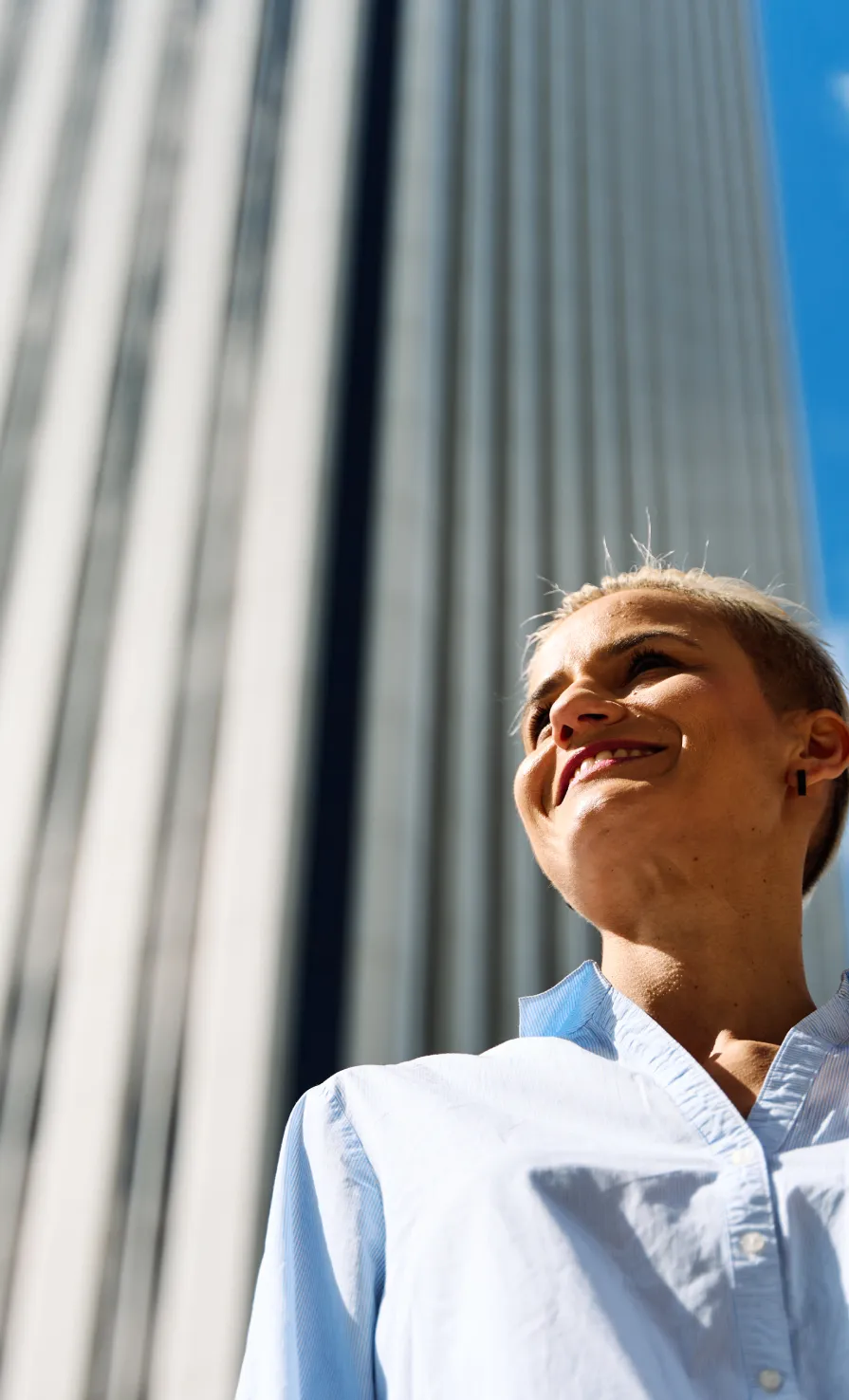 Smiling person with short hair in a light blue shirt standing outside near a tall building.