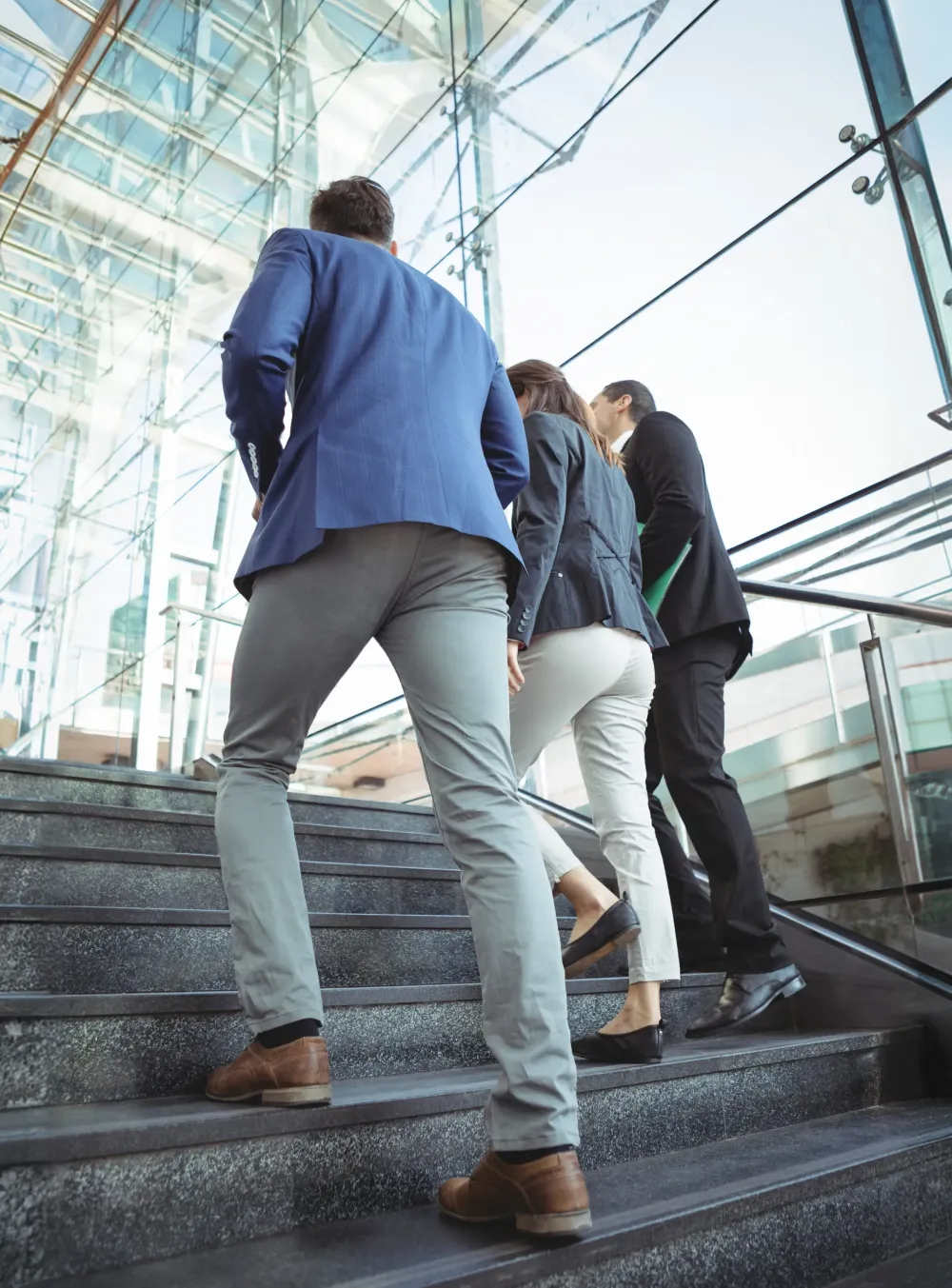 Three coworkers dressed in business attire walking up stairs inside a modern glass office building.