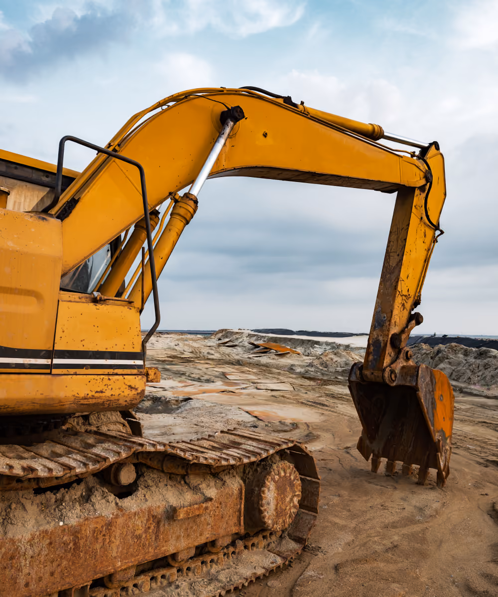 A yellow excavator with a rusted track is parked on a sandy construction site under a cloudy sky.