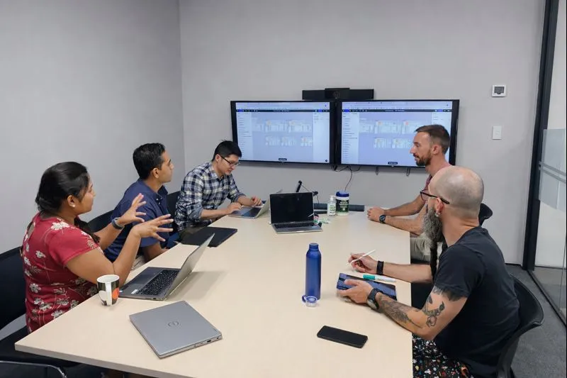 Team workshop around a meeting table, reviewing designs on two wall-mounted screens.