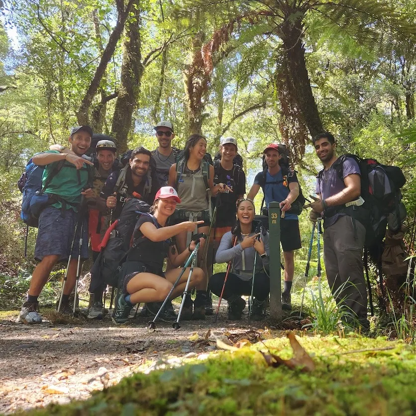 Group of hikers with backpacks and trekking poles posing beside a trail marker in New Zealand bush.