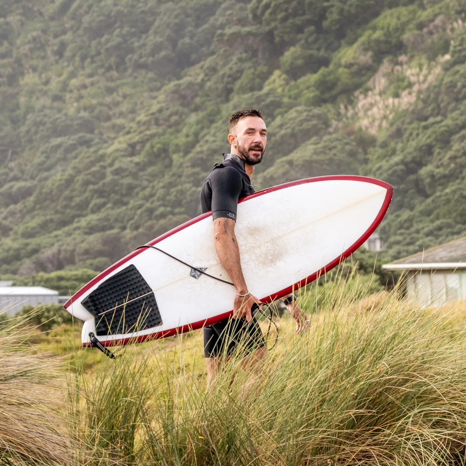 Man in a wetsuit carrying a white surfboard with a red edge walking through tall grass in front of a forested hill.