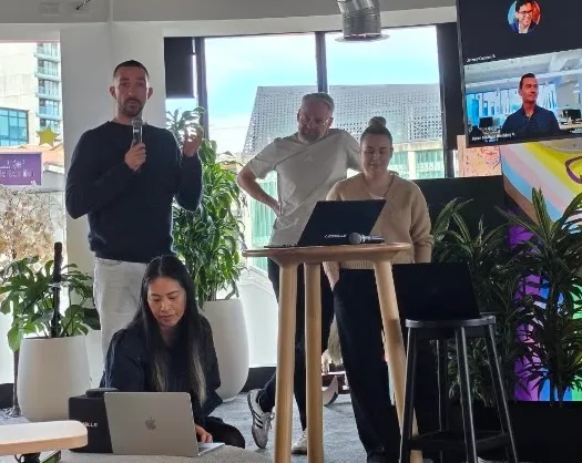 Four people in a modern office: one speaking with a microphone (Vlad), two at laptops, and one standing behind them.