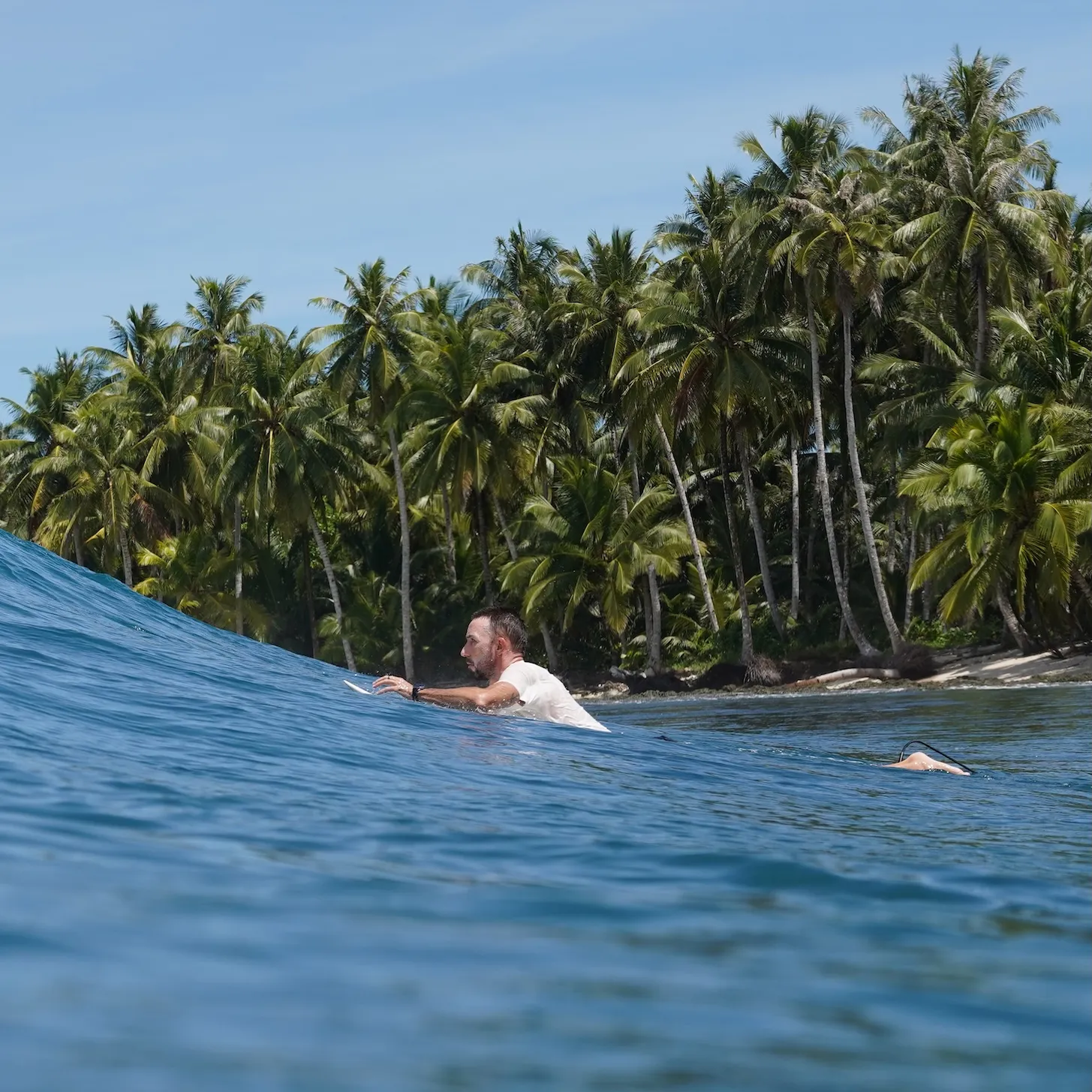 Vlad paddling on a surfboard in ocean water near a tropical island with tall palm trees.