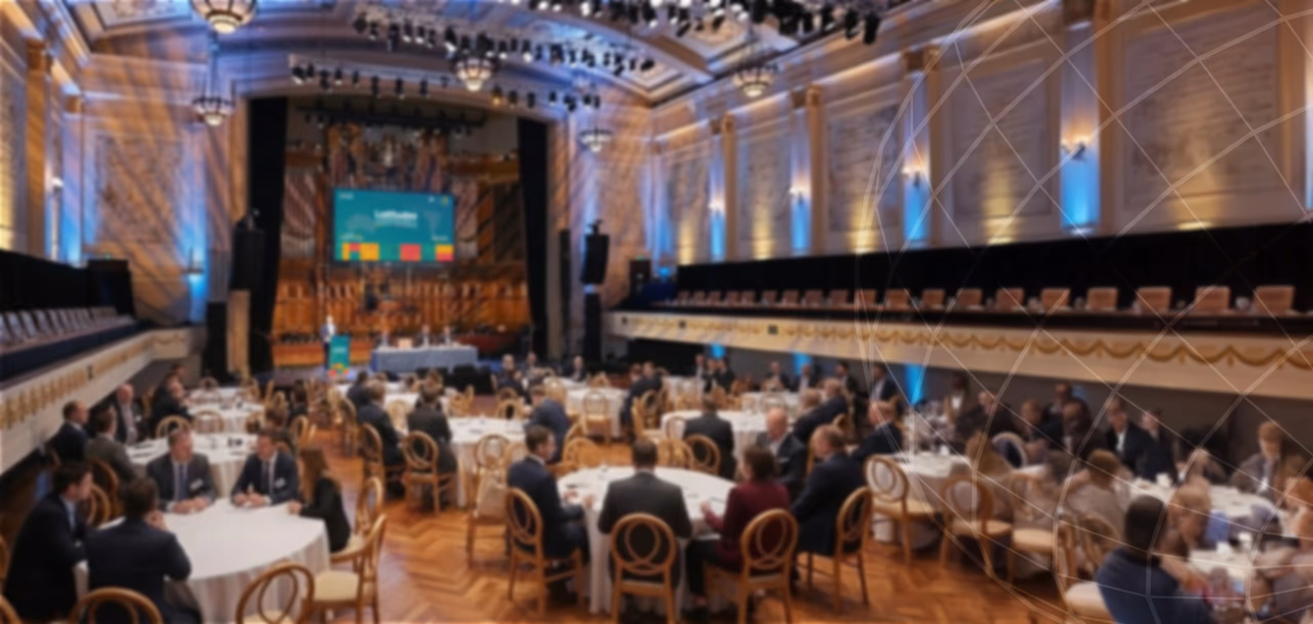Blurry image of a large conference room with round tables and people seated, facing a stage with a screen and blue uplighting on the walls.