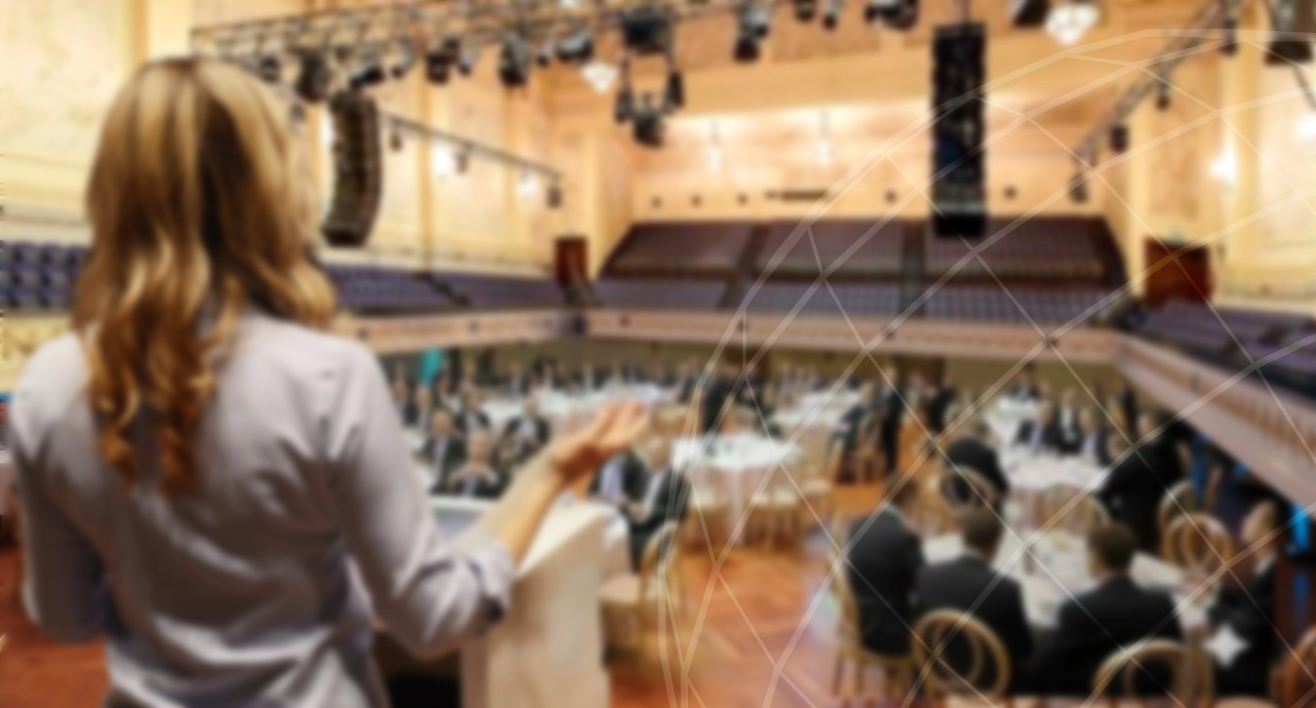 Speaker addressing a seated audience at a formal event in a large auditorium with round tables.