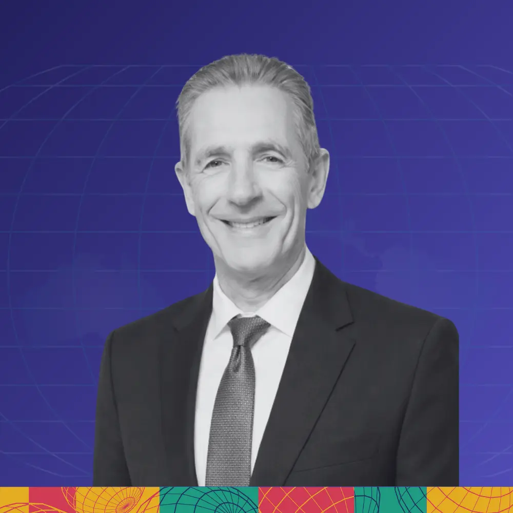 Smiling middle-aged woman wearing glasses, a collared shirt, and beaded necklaces, with a teal and green patterned background.