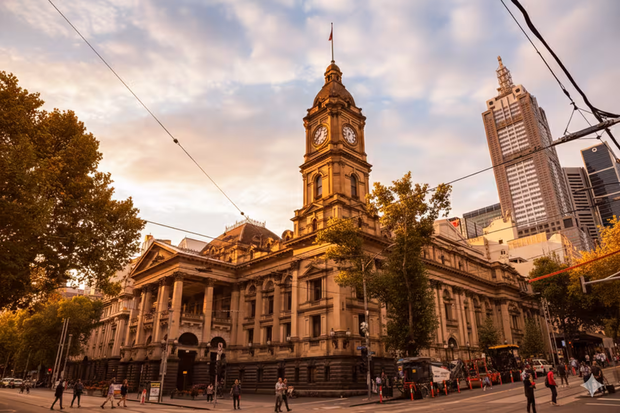 Historic Melbourne Town Hall building with clock tower at sunset, surrounded by trees and city skyscrapers.
