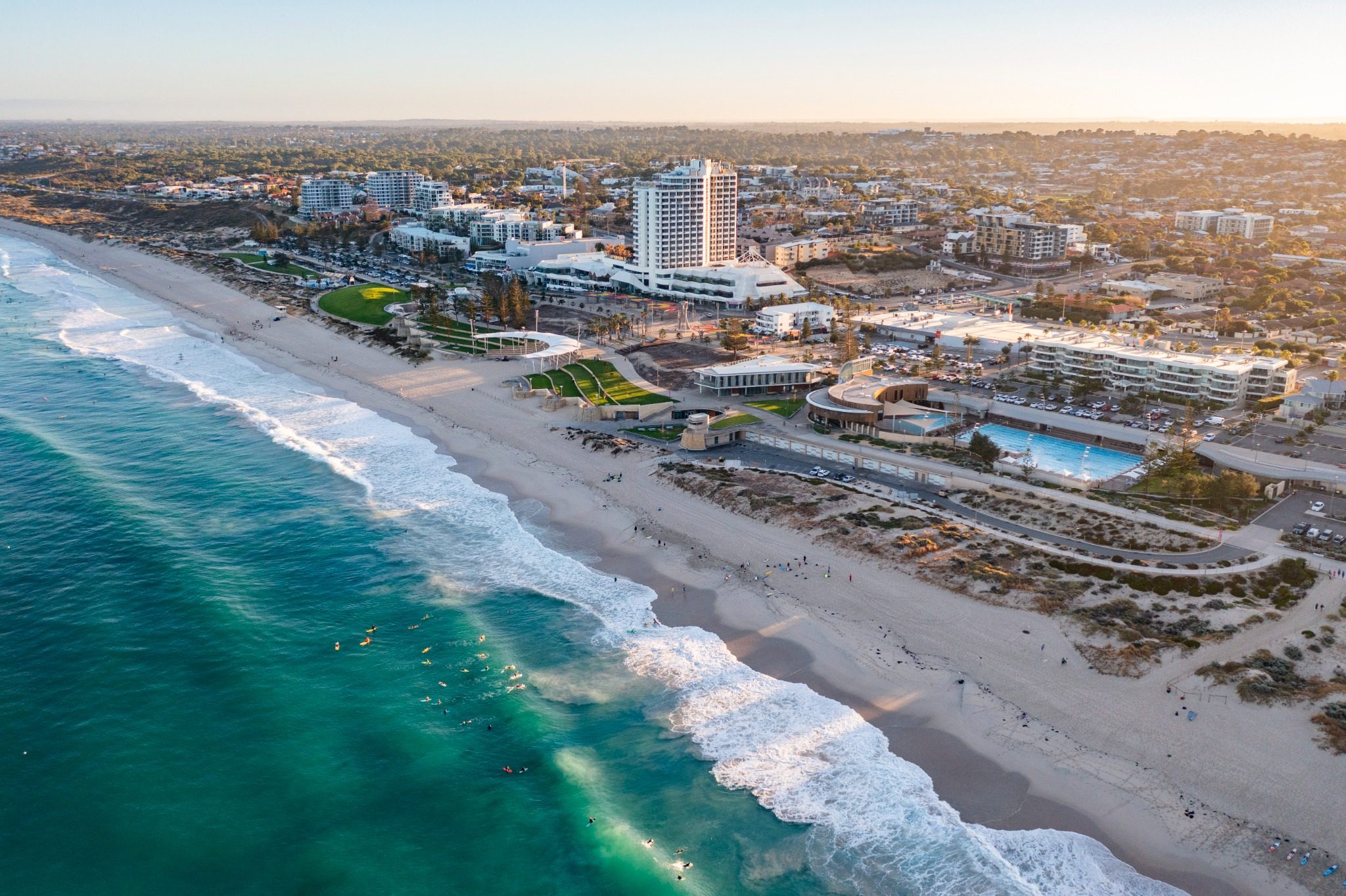 A coastal cityscape with a sandy beach, people surfing in the ocean, and buildings lining the shore at sunset.