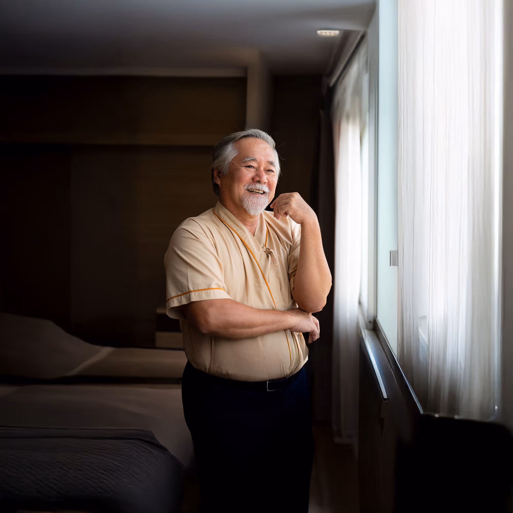 Smiling elderly man with gray hair and beard standing by a window with sheer curtains in a dimly lit room.