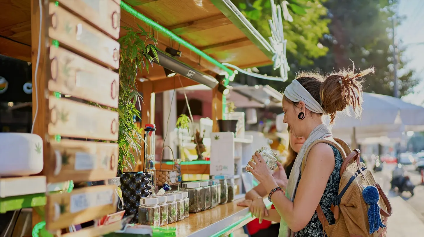 Woman with backpack examining jar of cannabis at an outdoor market stall with cannabis plants and products.