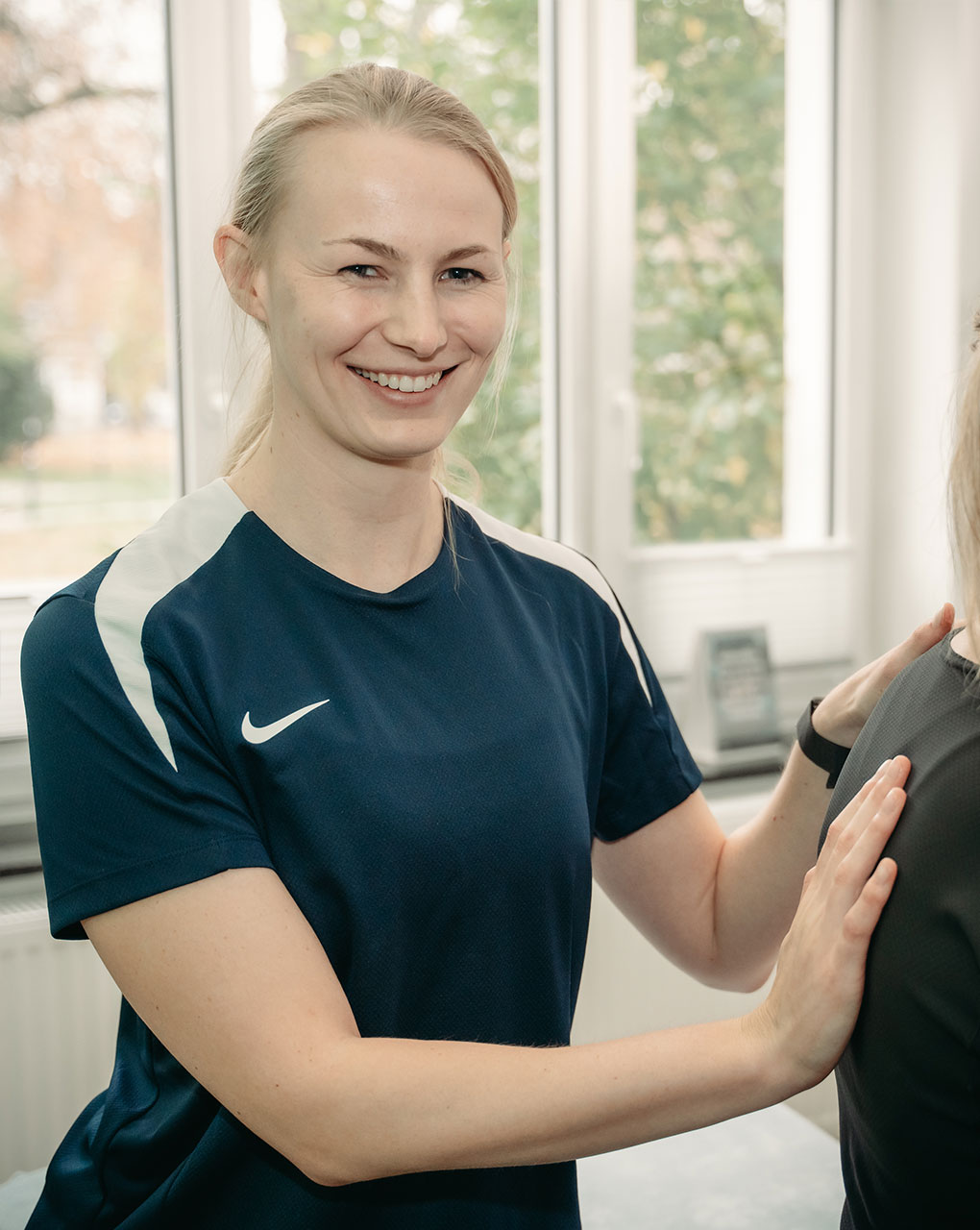 Physiotherapeutin in blauem Sportshirt lächelt und behandelt einen Patienten.
