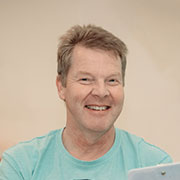 Lächelnder Mann mittleren Alters mit kurzem Haar und blauem T-Shirt vor hellbeigem Hintergrund.