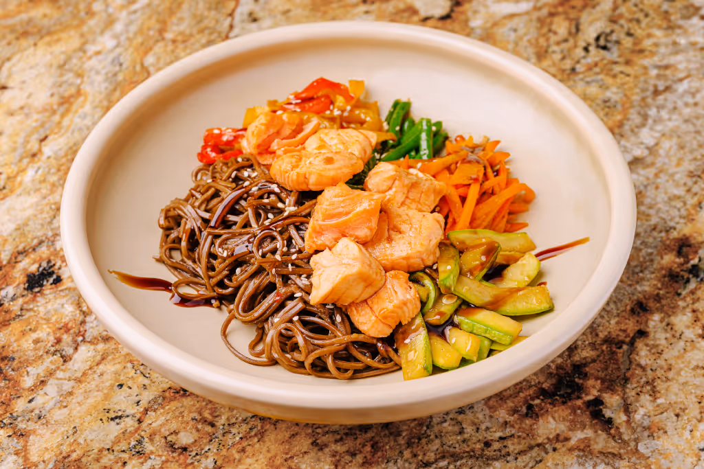 Bowl of soba noodles topped with cooked salmon chunks, sautéed zucchini, carrots, green beans, and red peppers, drizzled with sauce and sprinkled with sesame seeds.