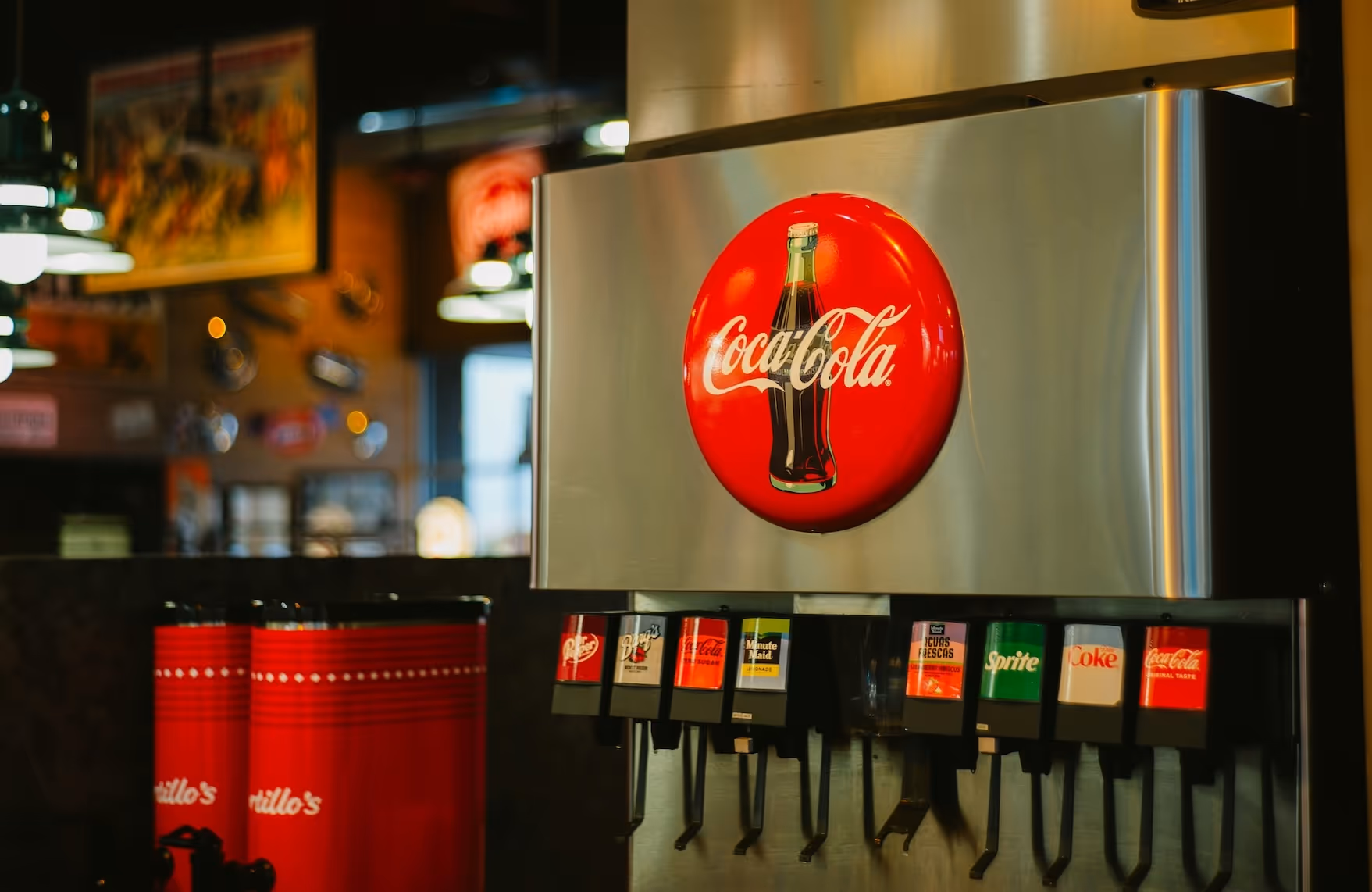 Soda fountain machine with Coca-Cola logo and multiple drink options including Dr Pepper, Diet Dr Pepper, Coca-Cola, Minute Maid, Sprite, and Coke, in a dimly lit restaurant setting.