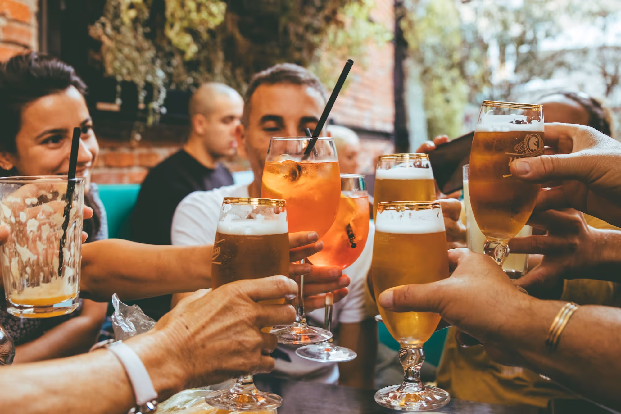 Group of friends clinking glasses of beer and cocktails in a lively outdoor setting.