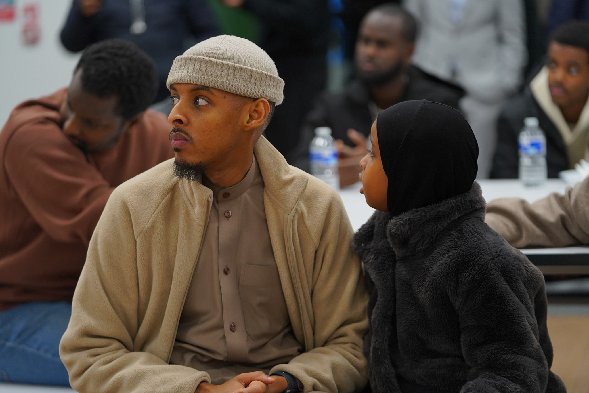 Man wearing a beige hat and jacket sitting next to a child in a black hooded coat, both looking to the left in a group setting.