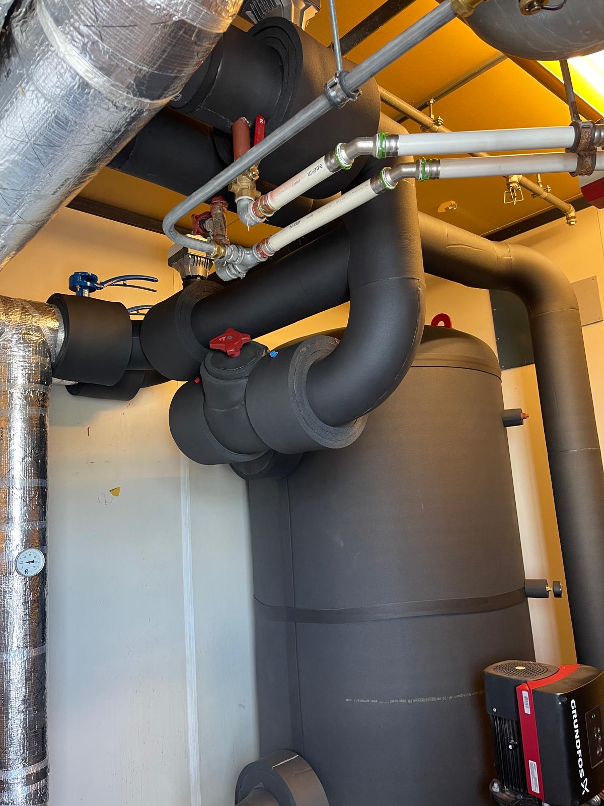 Zwarte Armaflex-isolatie op leidingwerk, bochten en afsluiters in een technische ruimte, met daaronder een geïsoleerd vat